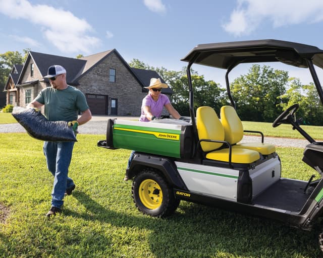 A man loads a bag of soil into the back of a John Deere Gator.