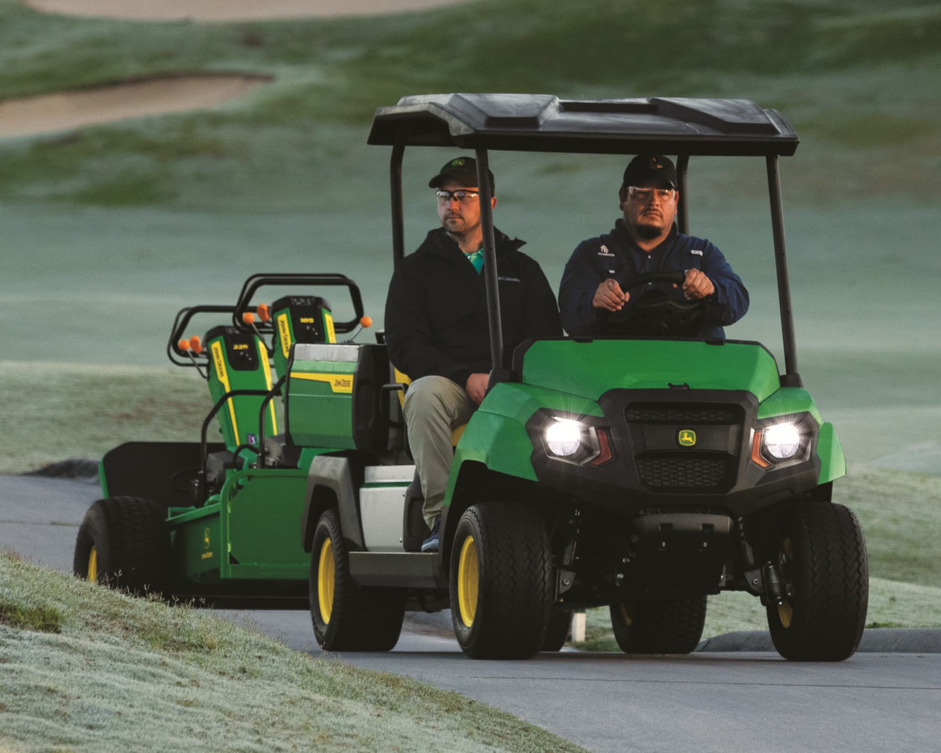 Two workers ride in a John Deere Gator on a golf course.