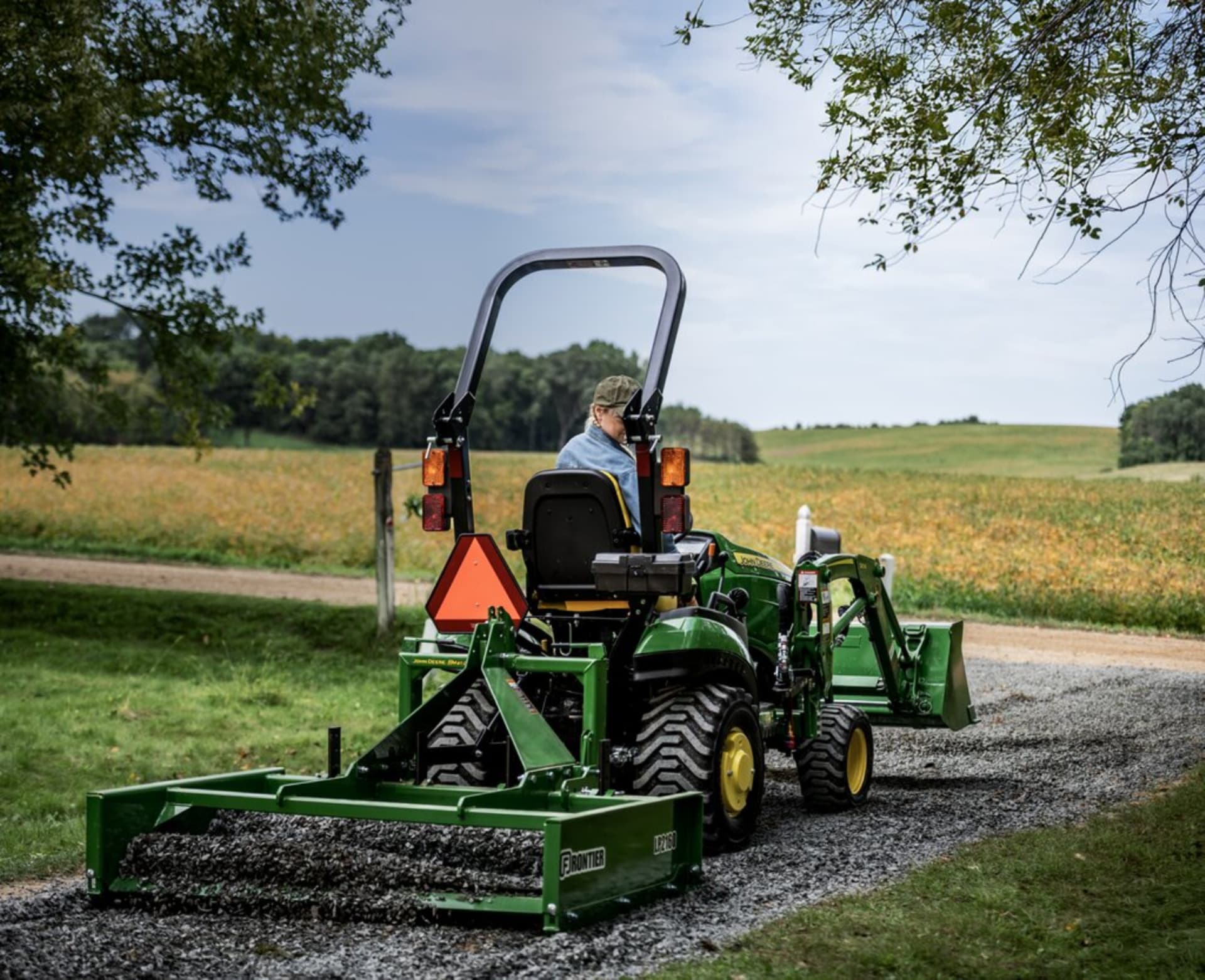 A John Deere 1025R with a grader attachment smooths a gravel driveway.